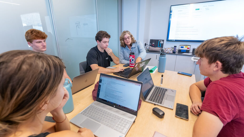 Students sitting around a table with their professor working on their laptops