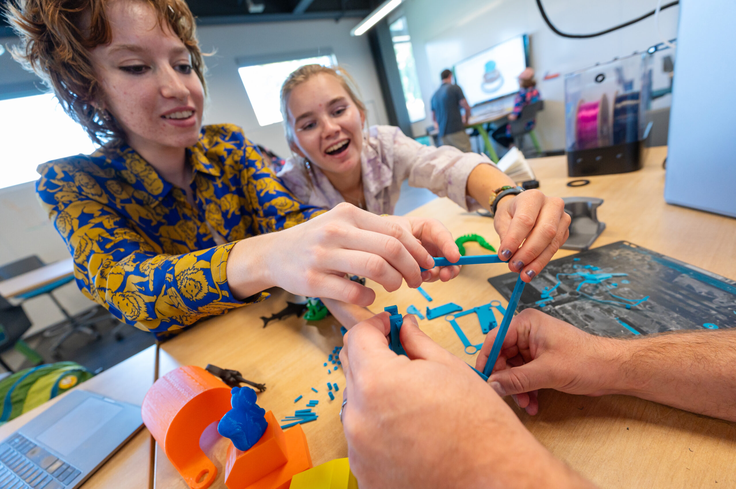 Two design engineering students smile as they assemble a 3D-printed model on a table, with other colorful parts and tools visible around them.