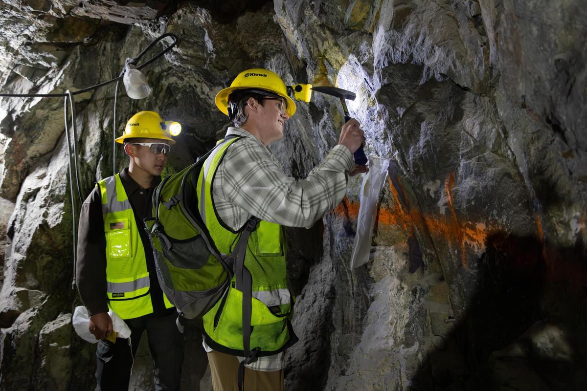 Two geological engineers in yellow hard hats and safety vests examine rock formations in a mine, using tools and equipment for analysis.
