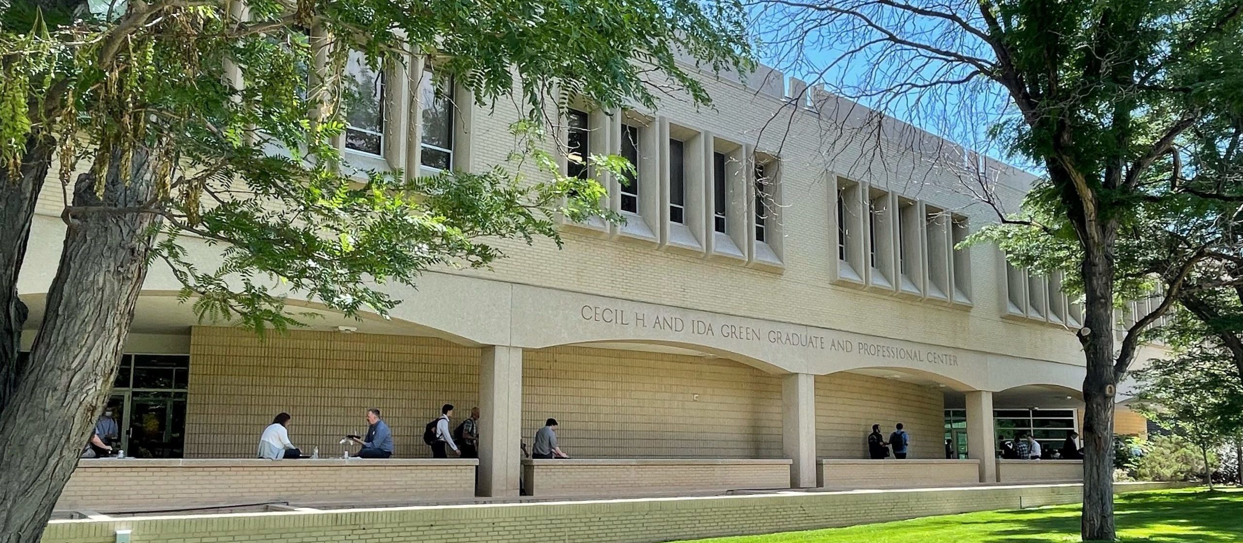 A light-colored two-story building behind a green lawn and several trees. There is a covered outdoor section on the first floor with people walking around, and the top floor has narrow rectangular windows in clusters of five. The words "Cecil H and Ida Green Graduate and Professional Center" are engraved on the awning above the first floor.