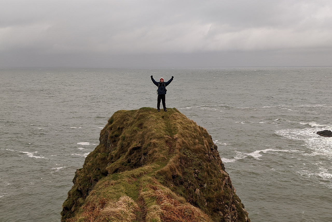 woman standing at the edge of a clip with sea below facing camera with her arms raised