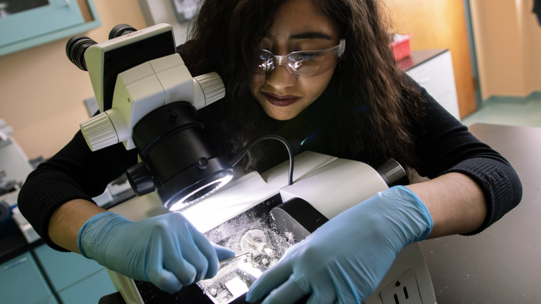 A woman in blue gloves looking through a microscope.