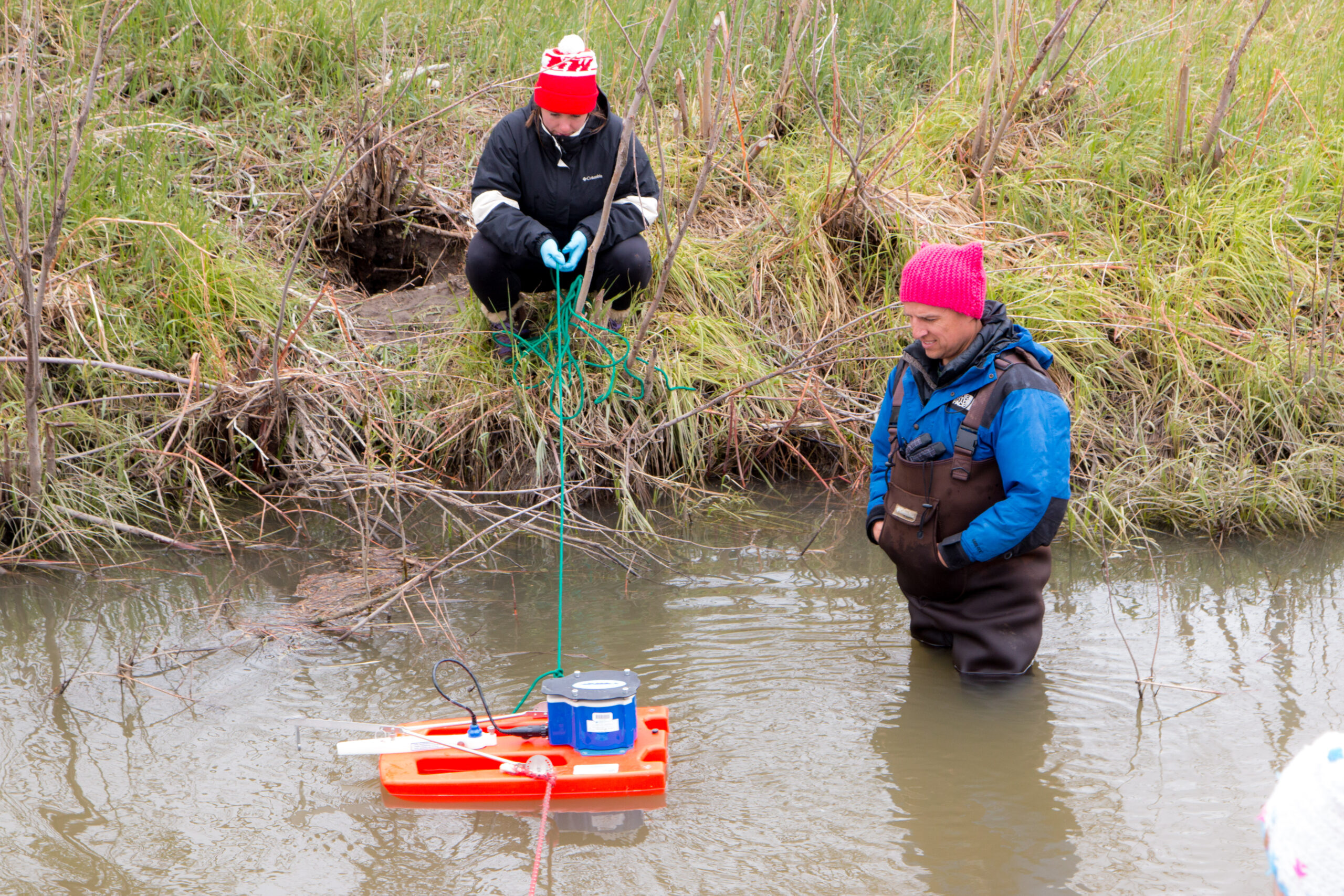 Two people, one on a river bank the other in the river with a floatation device collecting water.