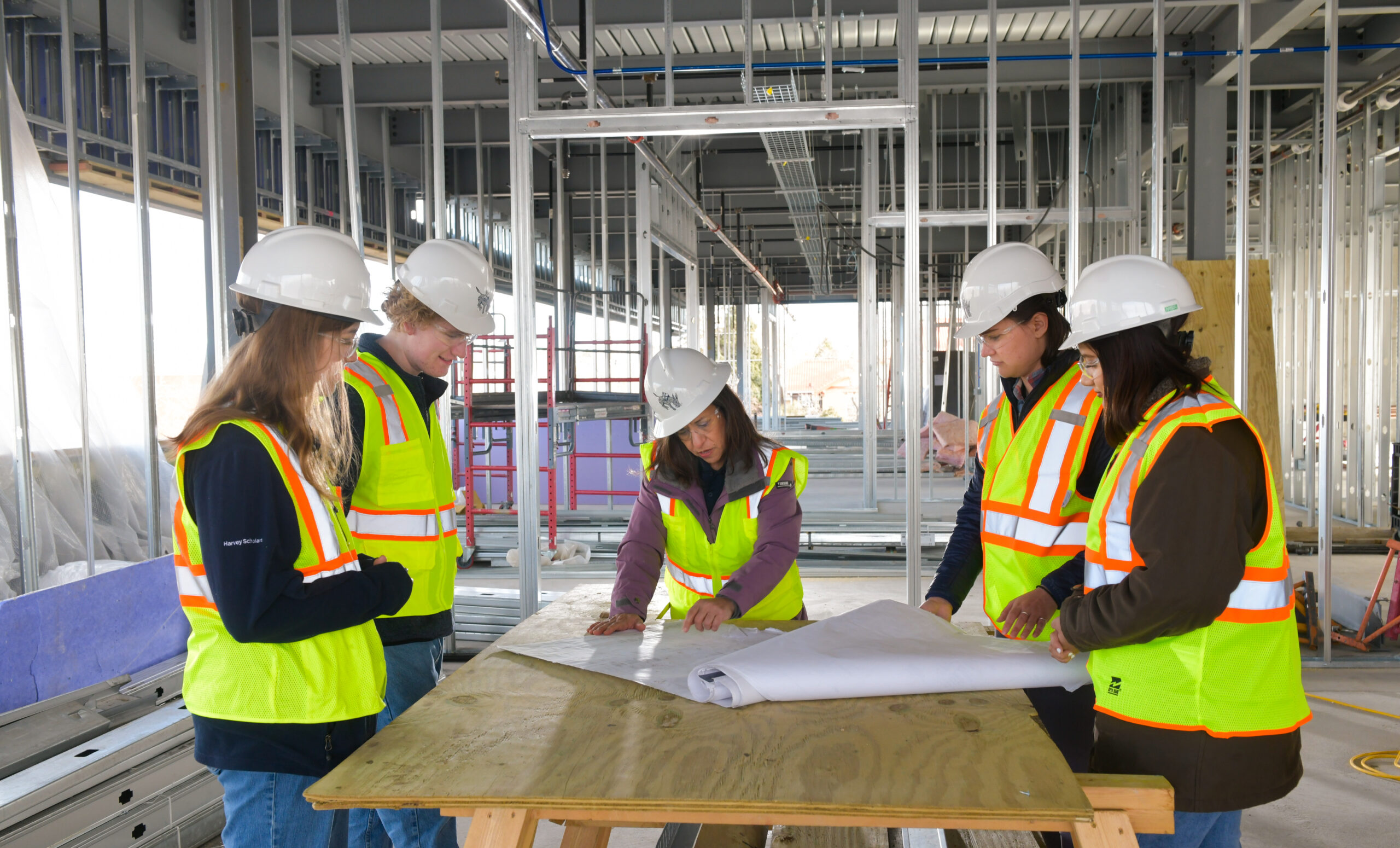 Professor at a table in a construction zone pointing out something on a plan to four students.