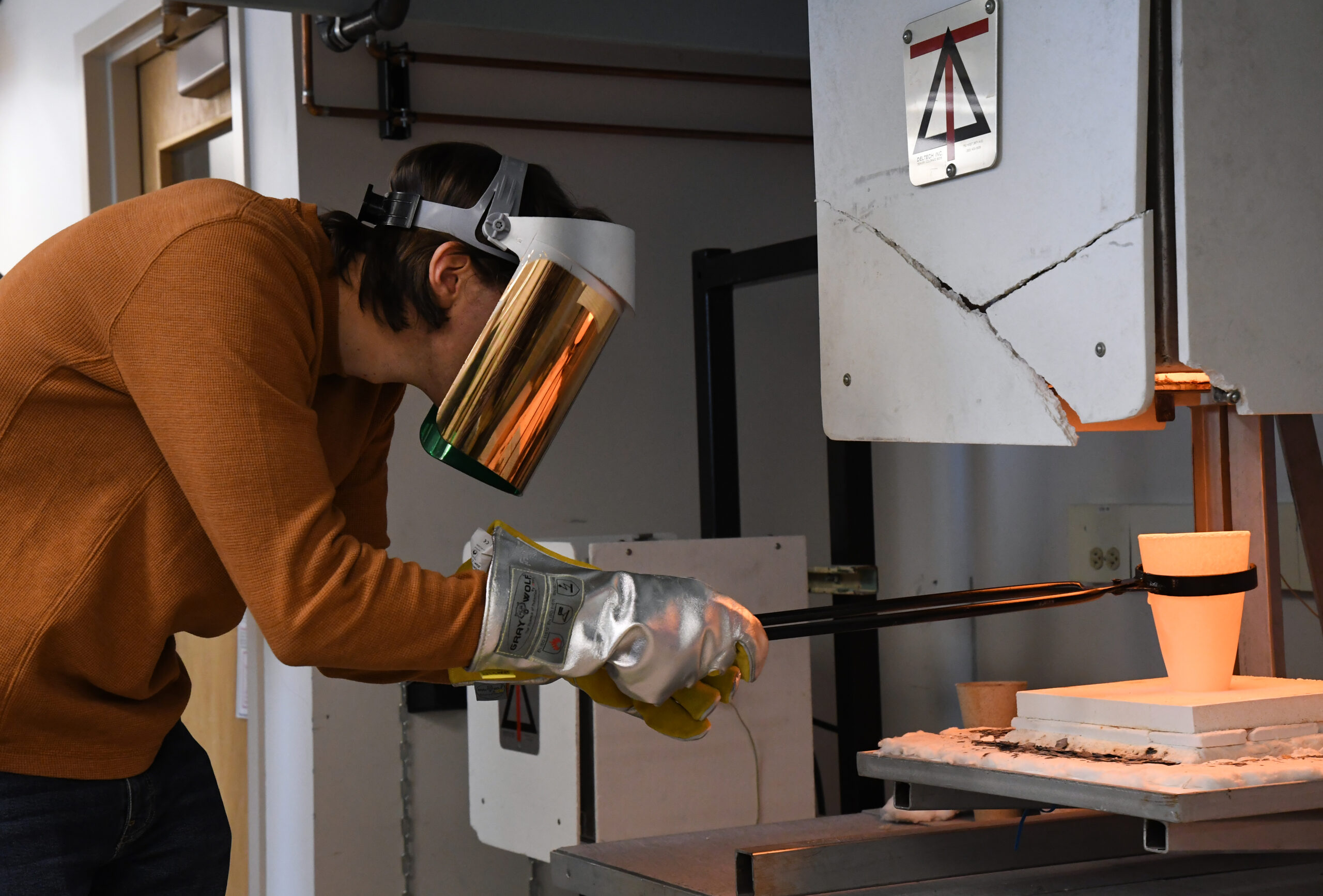 Student in a heat shield mask collects something in a hot cup held with tongs.