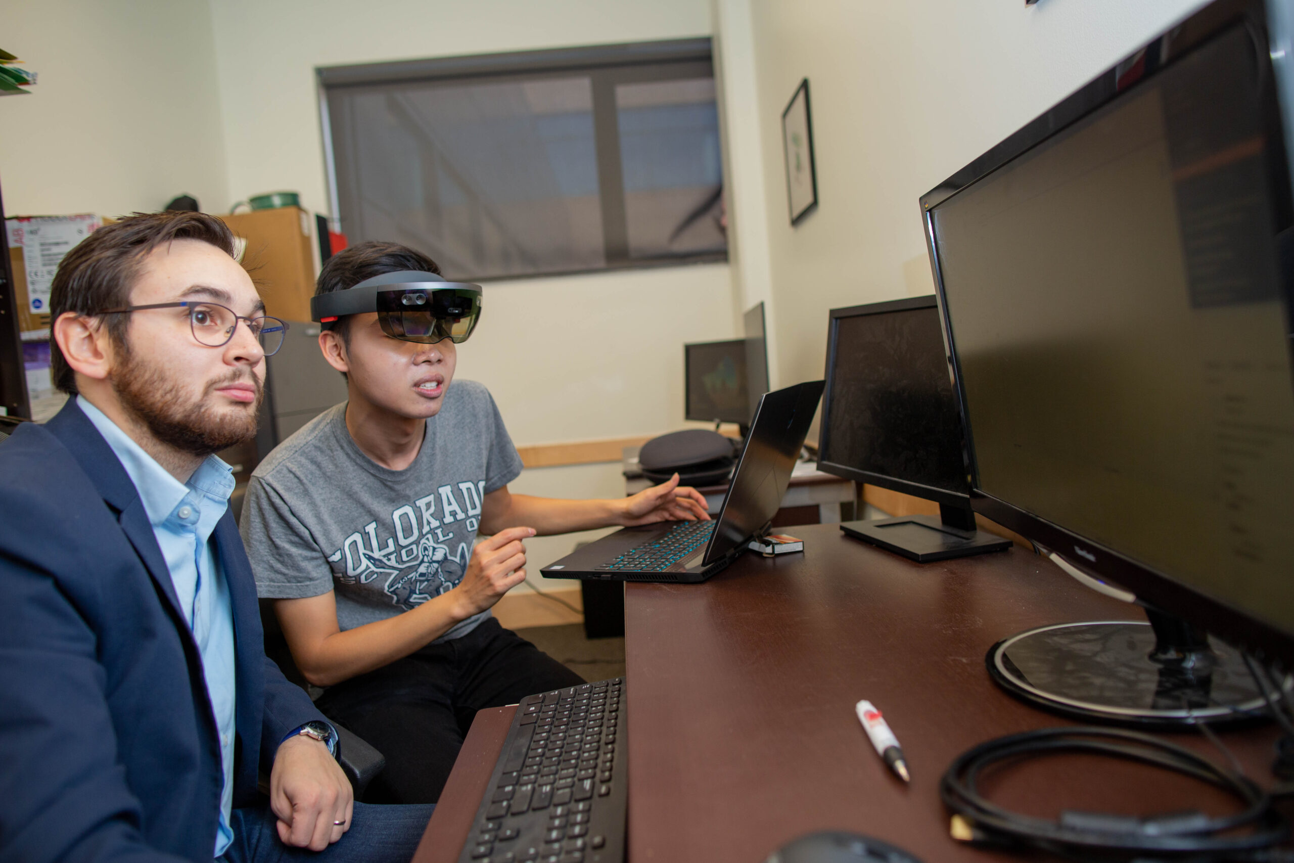 Two students looking at a computer, one with a set of goggles with a screen on them. 