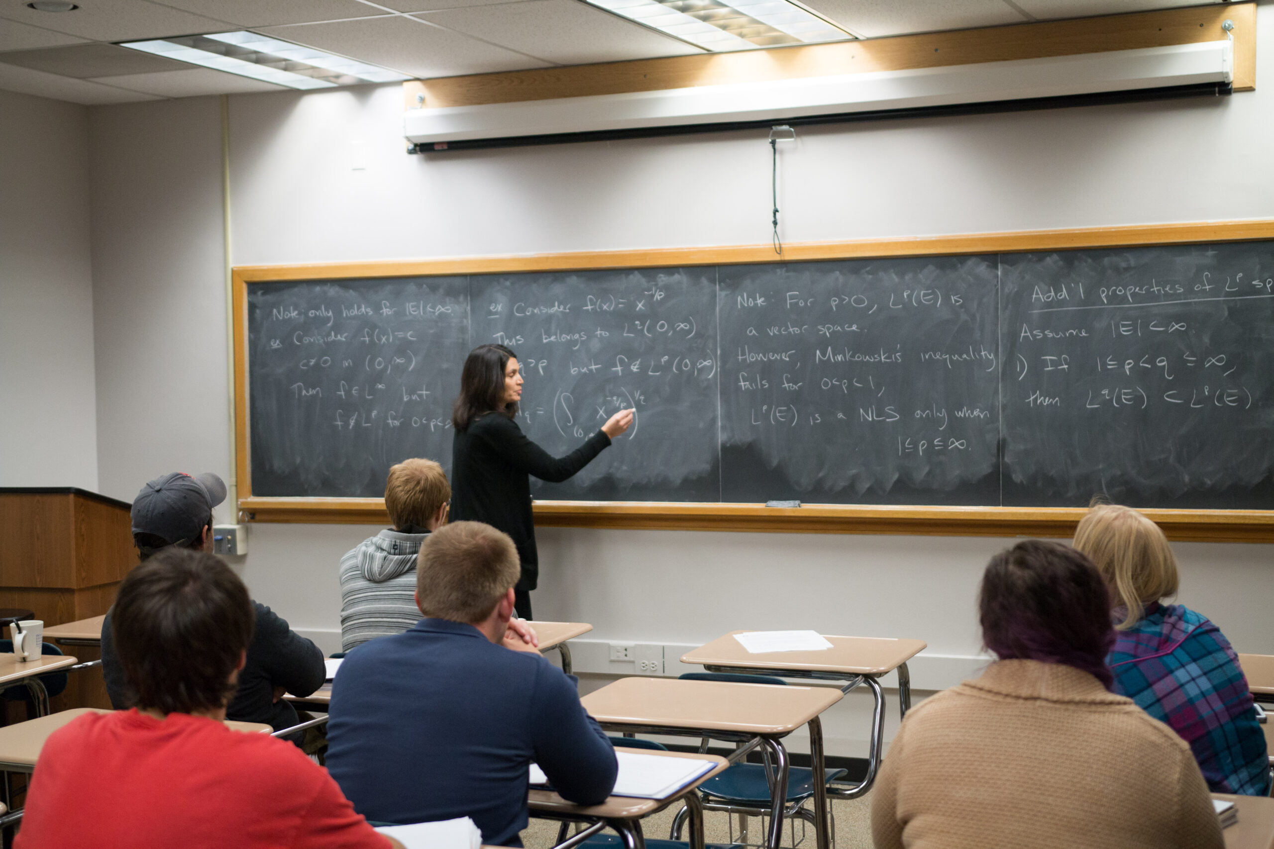 Professor at chalkboard writing out mathematical equations while students look on. 