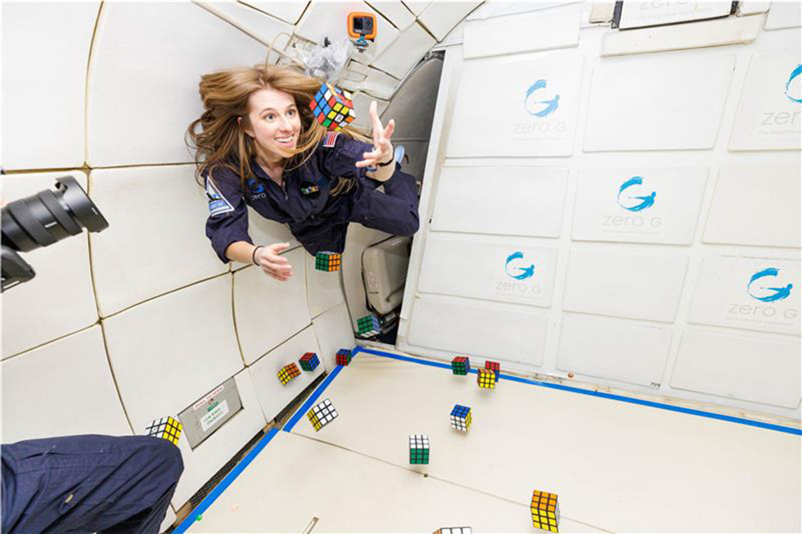 Bailey Burns in a zero-gravity environment reaches for floating Rubik's cubes amidst the interior of a spacecraft.